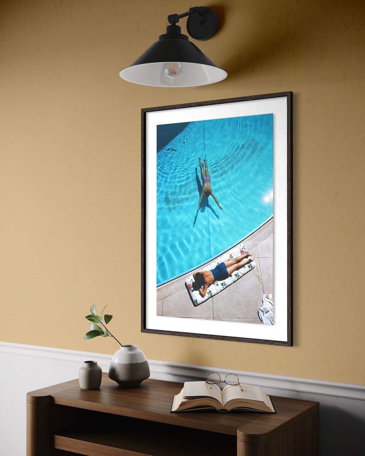 A framed art print depicting a person swimming in a bright blue pool, styled on a wooden console table against a warm beige wall under a black wall sconce. The Picturalist Slim Aarons Collection