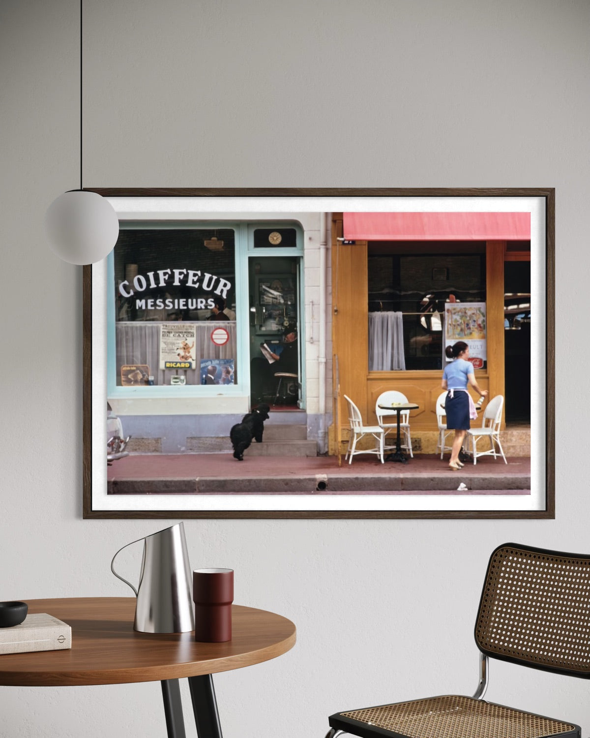 A framed art print depicting a Parisian street scene with a coffee shop and outdoor seating, displayed on a light gray wall above a wooden table with a lamp and chair. The Picturalist Slim Aarons Collection.