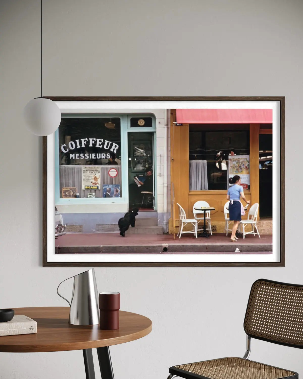 A framed art print depicting a Parisian street scene with a coffee shop and outdoor seating, displayed on a light gray wall above a wooden table with a lamp and chair. The Picturalist Slim Aarons Collection.