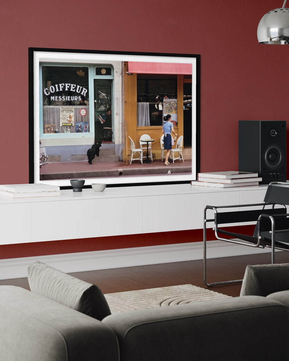A framed art print depicting a street scene with a coffee shop storefront and a person walking, displayed on a white wall above a console table in a modern living room setting. The Picturalist Slim Aarons Collection.