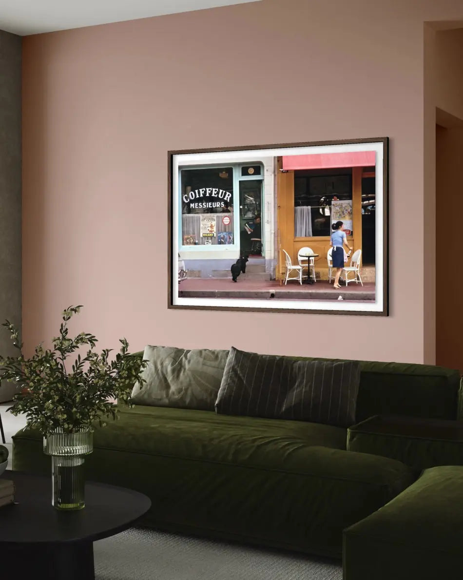A framed art print depicting a Parisian street scene with a cafe and a person, displayed on a dusty pink wall above a dark green velvet sofa in a modern living room setting. The Picturalist Slim Aarons Collection.