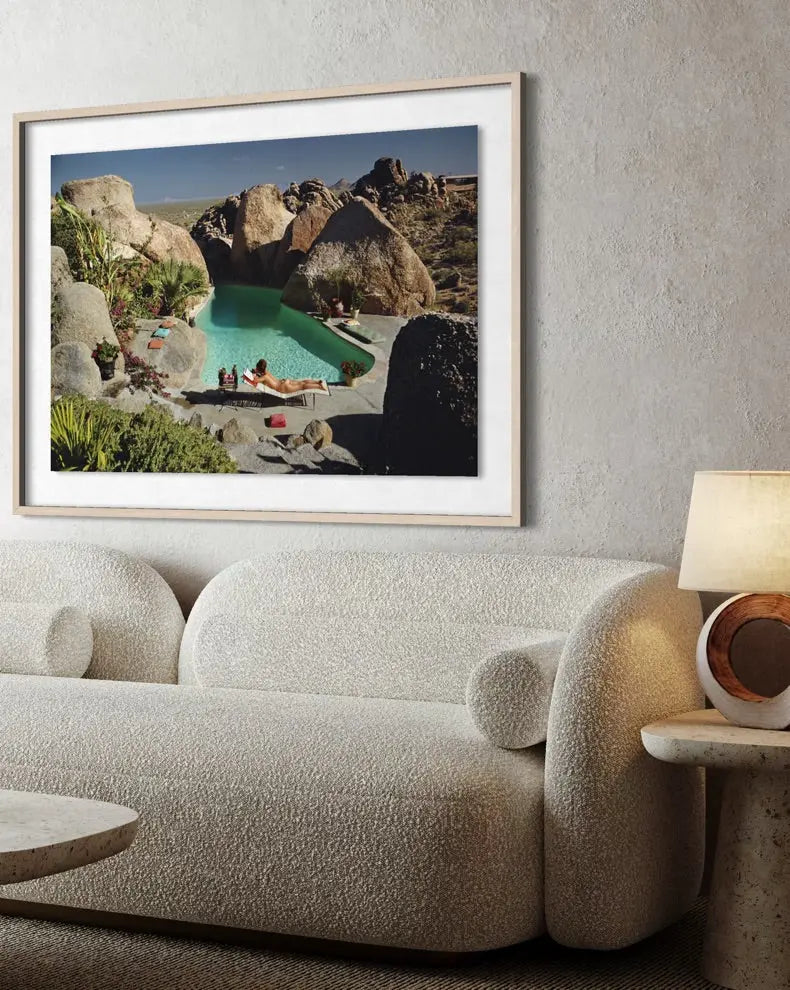 A framed art print depicting a scenic desert pool surrounded by large rocks, displayed on a textured light gray wall above a modern beige boucle sofa with a lamp on a side table. The Picturalist Slim Aarons Collection.