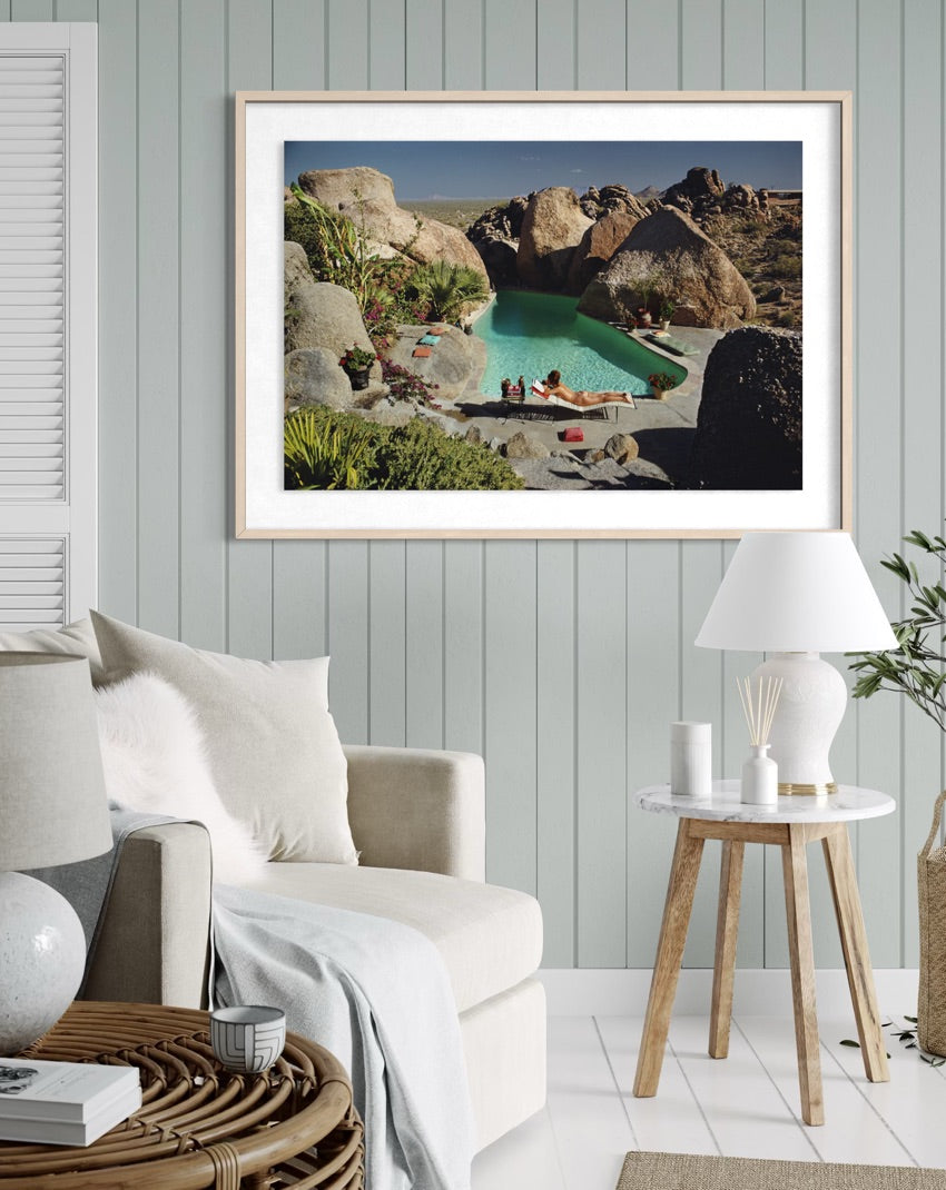 A framed art print depicting a scenic desert poolside landscape with large boulders and a turquoise pool, styled in a modern living room setting above a beige sofa.