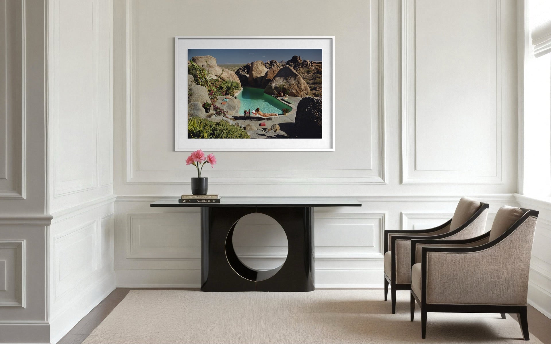 A framed landscape art print depicting a rocky coastal cove with turquoise water, displayed on a white paneled wall above a black console table in a modern living room setting.