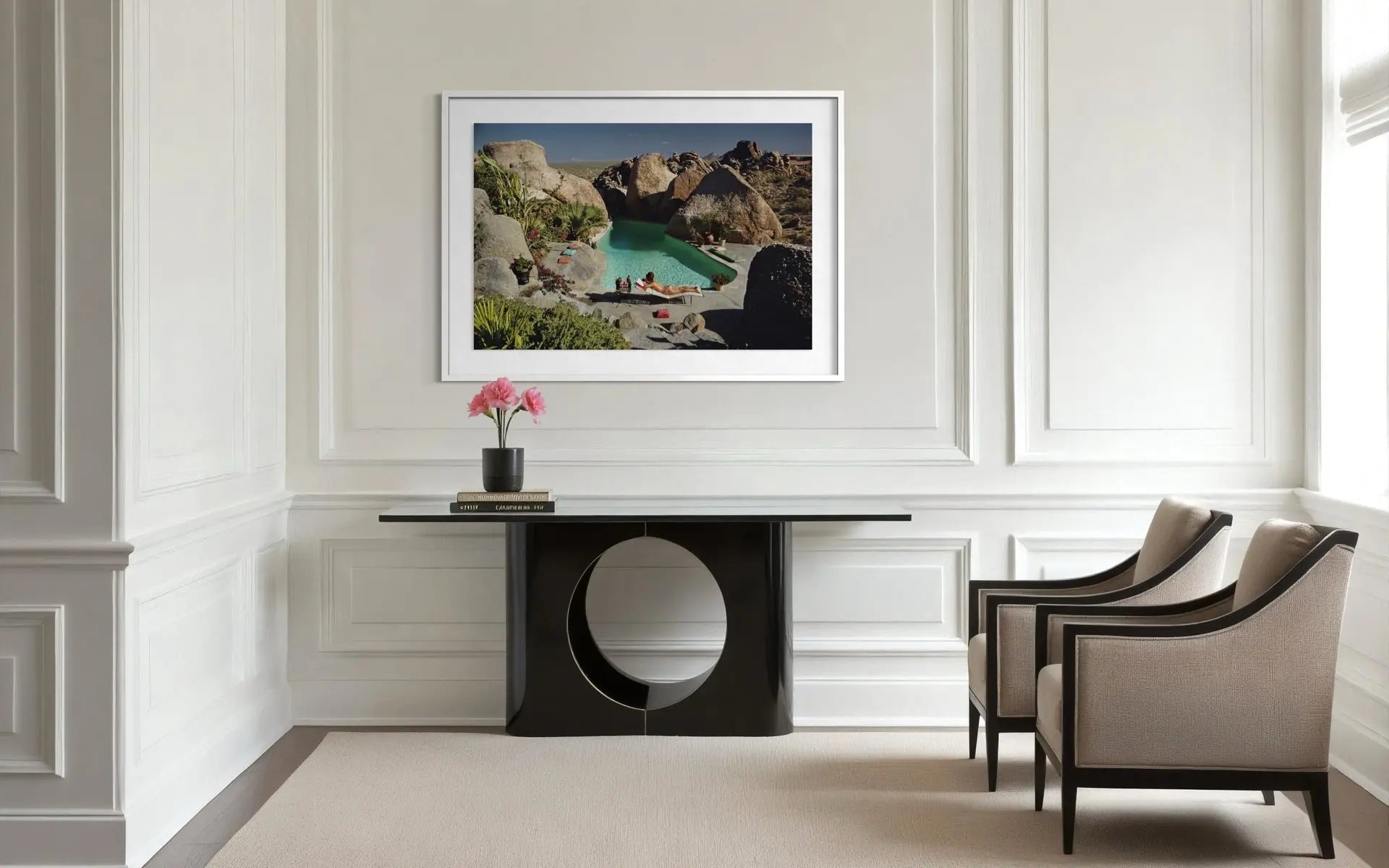 A framed landscape art print depicting a rocky coastal cove with turquoise water, displayed on a white paneled wall above a black console table in a modern living room setting.