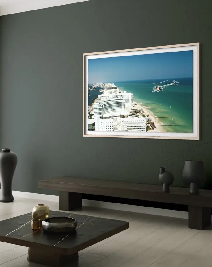 A framed photographic print of a coastal city and ocean scene hangs on a dark green wall above a long bench in a modern living room setting. The Picturalist Slim Aarons Collection.