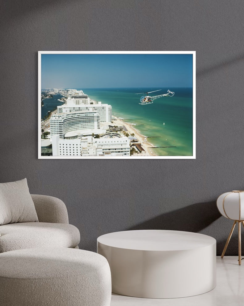 A framed coastal photograph print hanging on a dark gray wall above a modern white round coffee table and light beige upholstered seating. The print shows a bright blue sky, turquoise ocean, beach, and white buildings. The Picturalist Slim Aarons Collection.