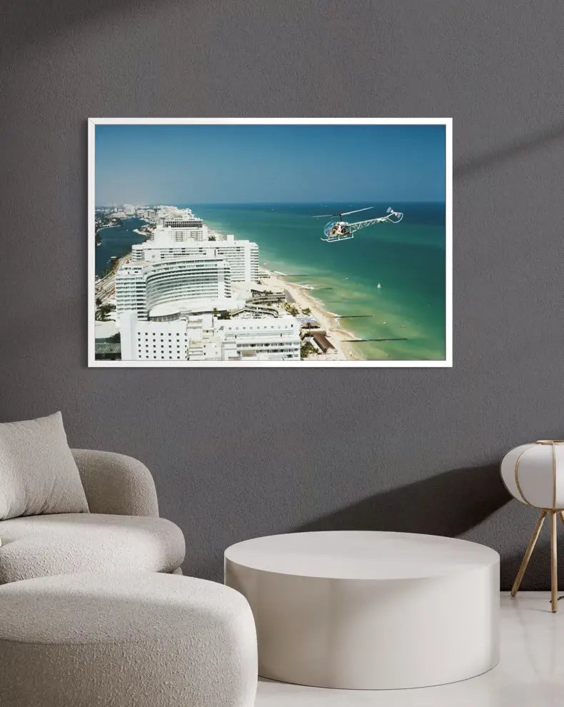 A framed coastal photograph print hanging on a dark gray wall above a modern white round coffee table and light beige upholstered seating. The print shows a bright blue sky, turquoise ocean, beach, and white buildings. The Picturalist Slim Aarons Collection.