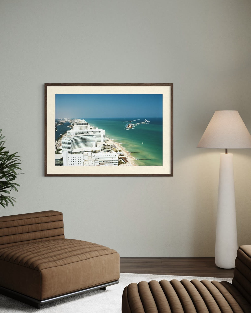 A framed photographic print of a coastal resort scene with turquoise ocean, white buildings, and a pier, displayed on a light gray wall above a brown upholstered lounge chair in a modern living room. The Picturalist Slim Aarons Collection.