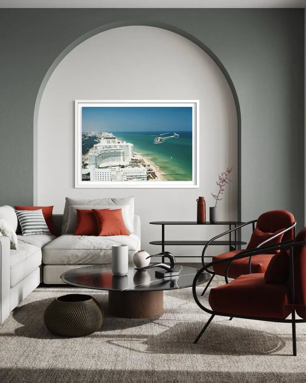 A framed art print depicting a coastal cruise ship scene in turquoise water, displayed on a gray wall in a modern living room setting with a white sofa, red accent pillows, and a red armchair. The Picturalist Slim Aarons Collection.