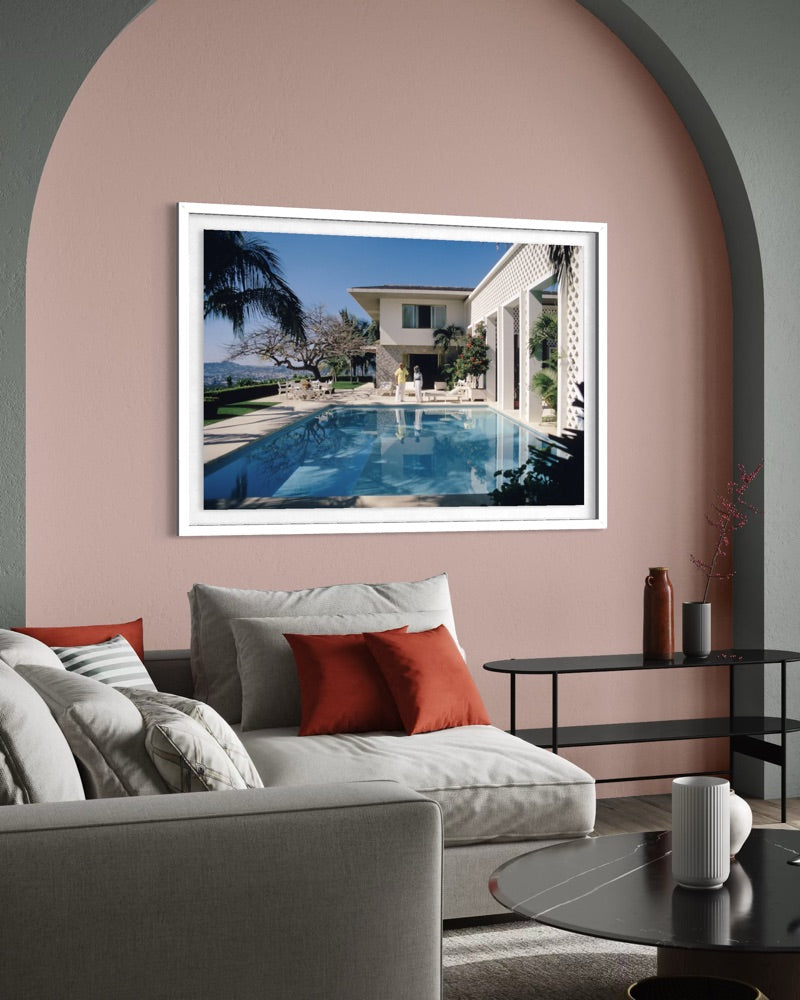 A framed art print depicting a luxurious villa with a swimming pool, displayed on a dusty pink wall in a modern living room setting. The Picturalist Wall Art and Photography. Slim Aarons Collection.