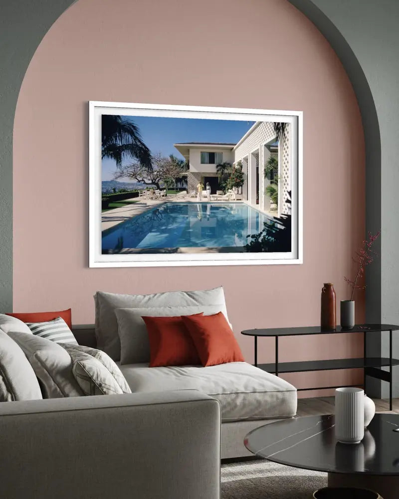 A framed art print depicting a luxurious villa with a swimming pool, displayed on a dusty pink wall in a modern living room setting. The Picturalist Wall Art and Photography. Slim Aarons Collection.