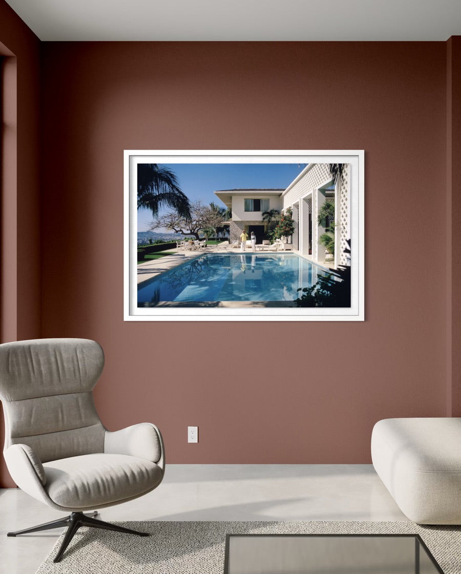 A framed wall art print depicting a luxurious outdoor poolside scene with a modern house, palm tree, and blue sky, displayed on a terracotta-colored wall in a styled living room setting. The Picturalist Wall Art and Photography. Slim Aarons Collection.
