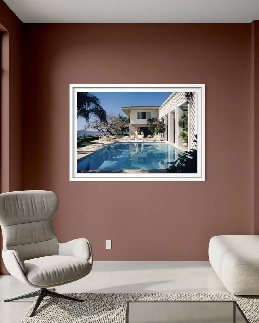 A framed wall art print depicting a luxurious outdoor poolside scene with a modern house, palm tree, and blue sky, displayed on a terracotta-colored wall in a styled living room setting. The Picturalist Wall Art and Photography. Slim Aarons Collection.