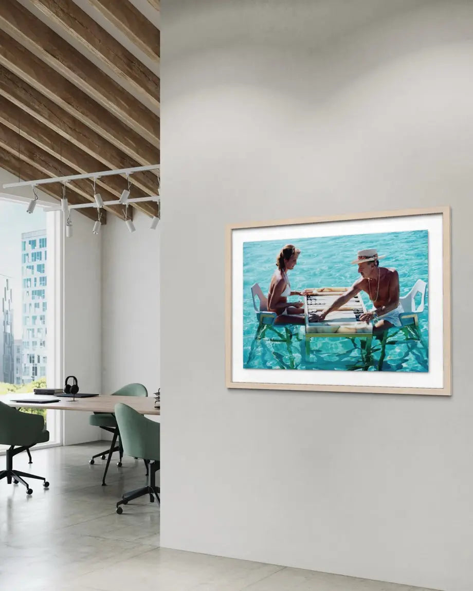 A framed art print depicting a couple sitting at a table by a turquoise ocean, displayed on a white wall in a modern office interior with wooden ceiling beams and office chairs. The Picturalist Wall Art and Photography. Slim Aarons Collection. Poolside.