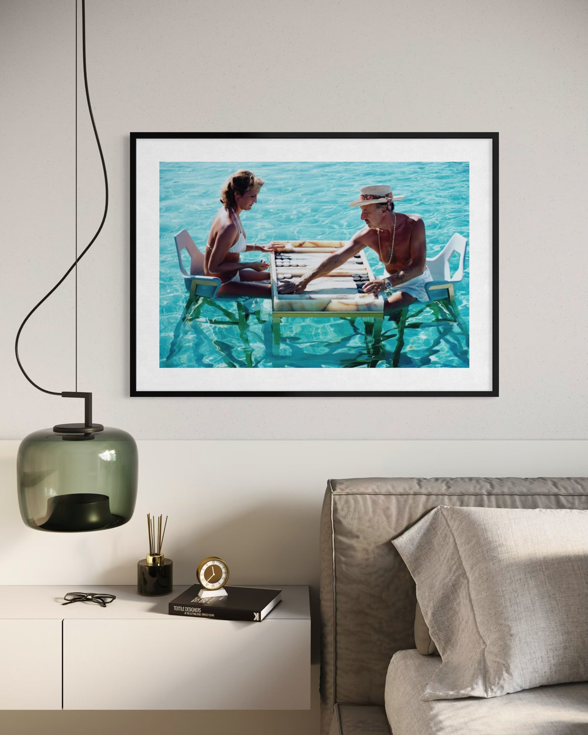 A framed art print depicting a couple sitting on liftable platforms in turquoise water, displayed on a light gray wall above a modern sideboard with a green pendant lamp and a gray sofa. The Picturalist Wall Art and Photography. Slim Aarons Collection. Poolside.