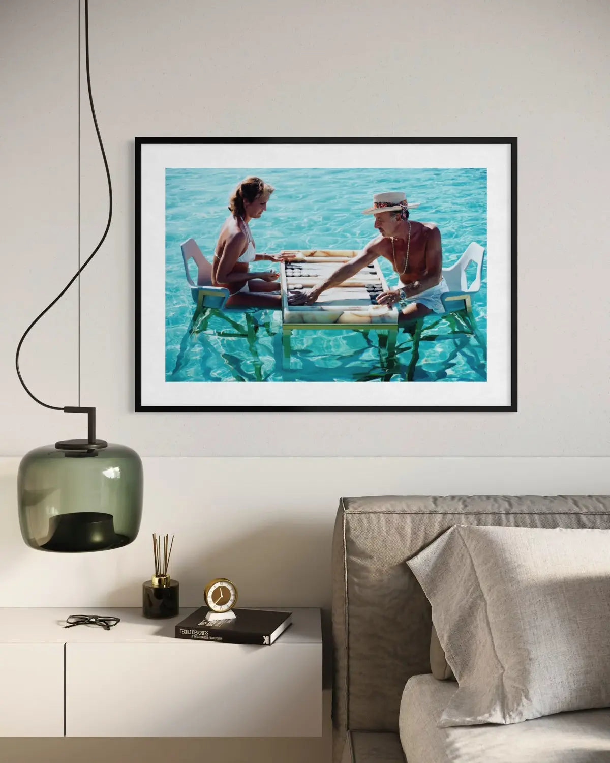 A framed art print depicting a couple sitting on liftable platforms in turquoise water, displayed on a light gray wall above a modern sideboard with a green pendant lamp and a gray sofa. The Picturalist Wall Art and Photography. Slim Aarons Collection. Poolside.