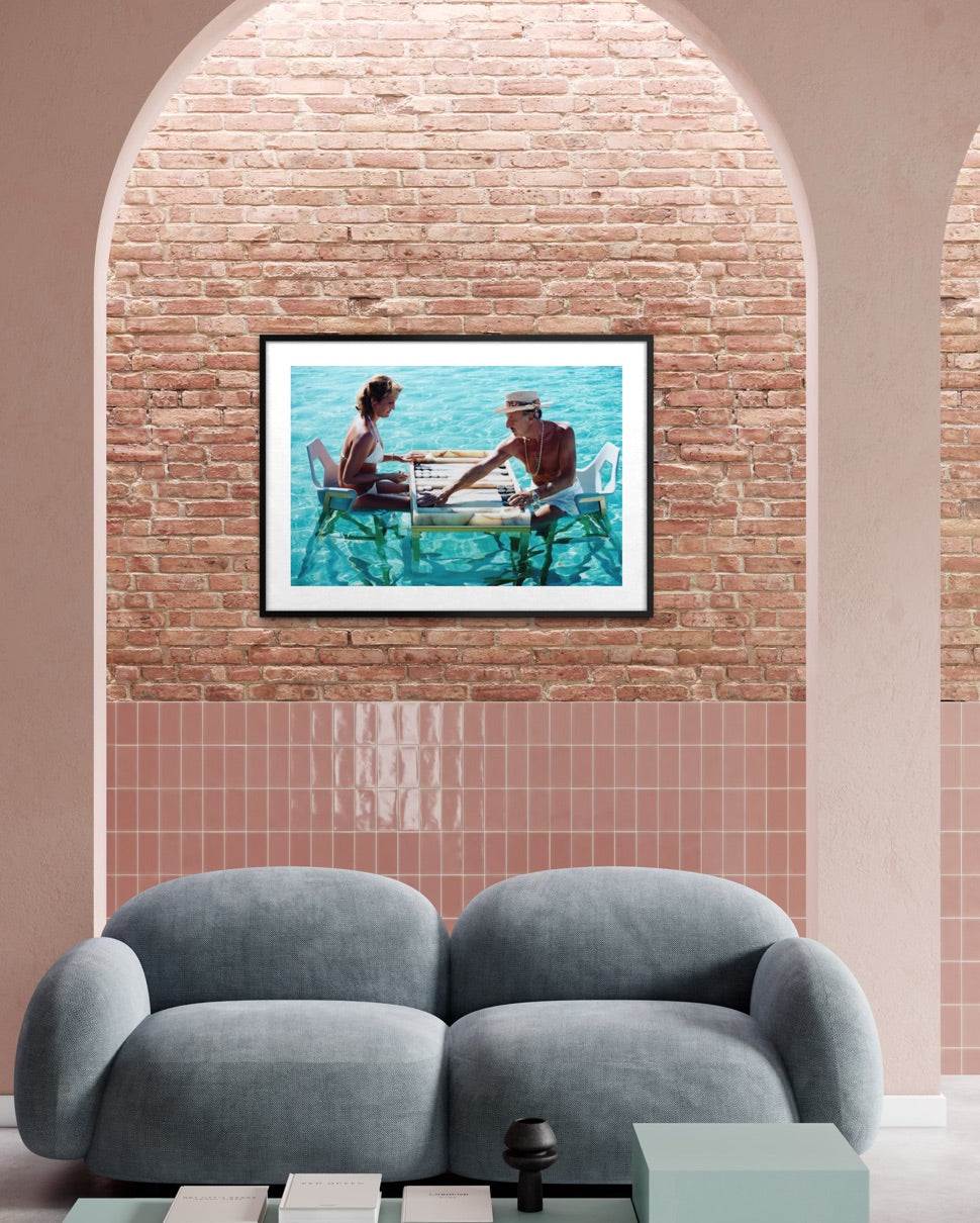 A framed art print depicting a couple sitting at a table by the sea, displayed on a brick and tile wall above a modern gray sofa in a living room setting. The Picturalist Wall Art and Photography. Slim Aarons Collection. Poolside.