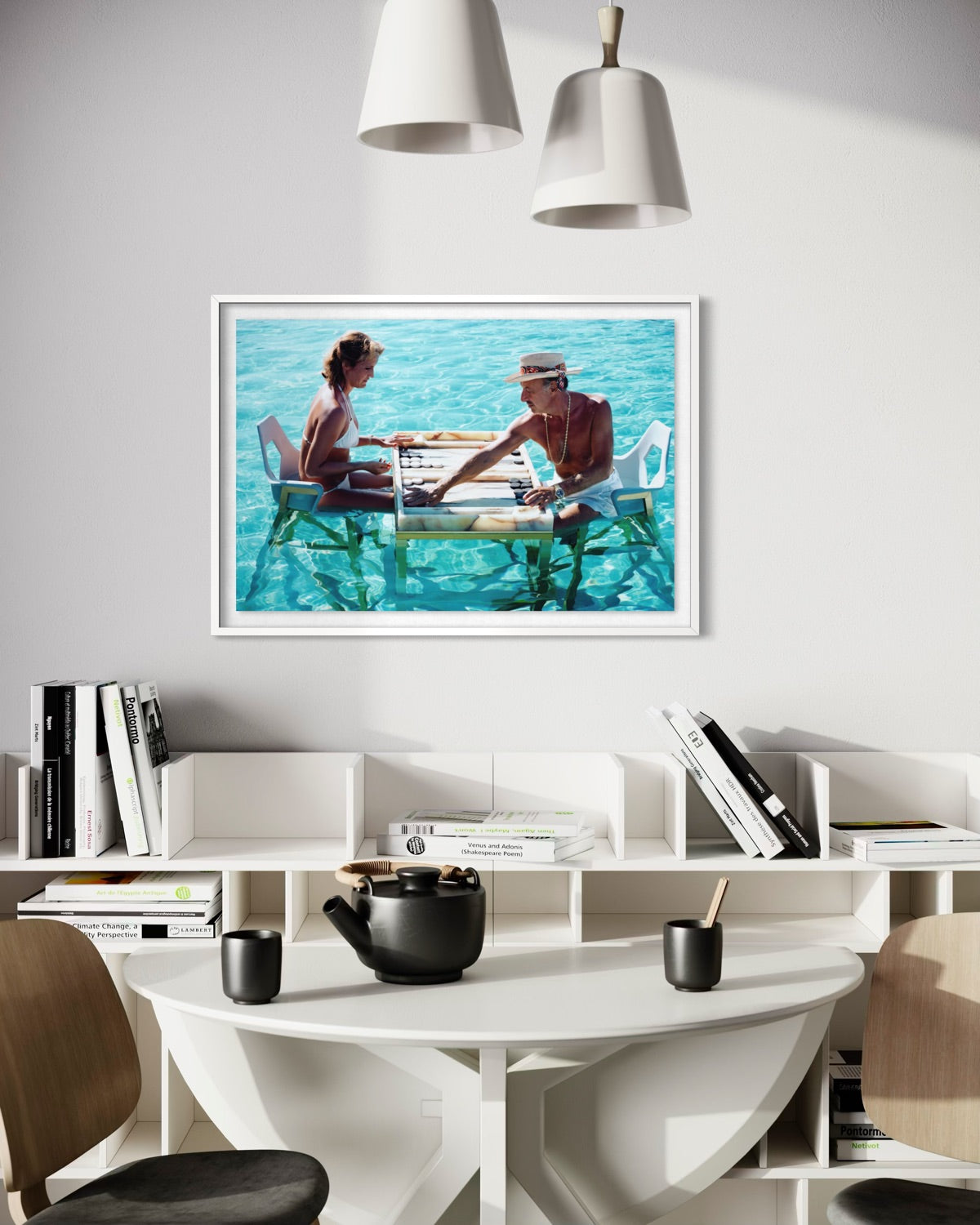 A framed art print depicting a couple playing with a game of cards on a wooden dock over turquoise water, displayed on a white wall above a modern white dining table and shelving unit. The Picturalist Wall Art and Photography. Slim Aarons Collection. Poolside.
