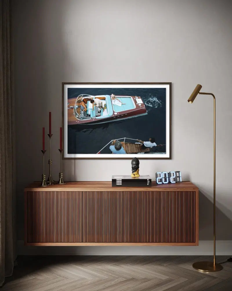 A framed wall art print showing an aerial view of a motorboat on dark water, displayed above a wooden console table in a modern living room setting with a floor lamp and decorative items. The Picturalist Wall Art and Photography for Designed Interiors. Slim Aarons Collection, Riva Boat. Getty Images Gallery.