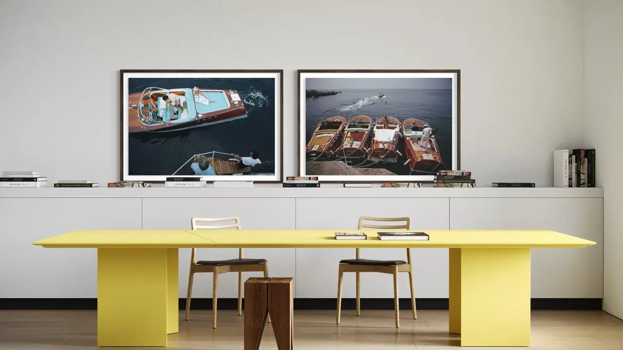A modern interior scene with a bright yellow rectangular table and chairs in front of a white wall, featuring two large framed photographic prints above. The prints depict boats at sea, one with people visible and one with boats docked. The Picturalist Wall Art and Photography for Designed Interiors. Slim Aarons Collection,Riva Boats. Getty Images Gallery.