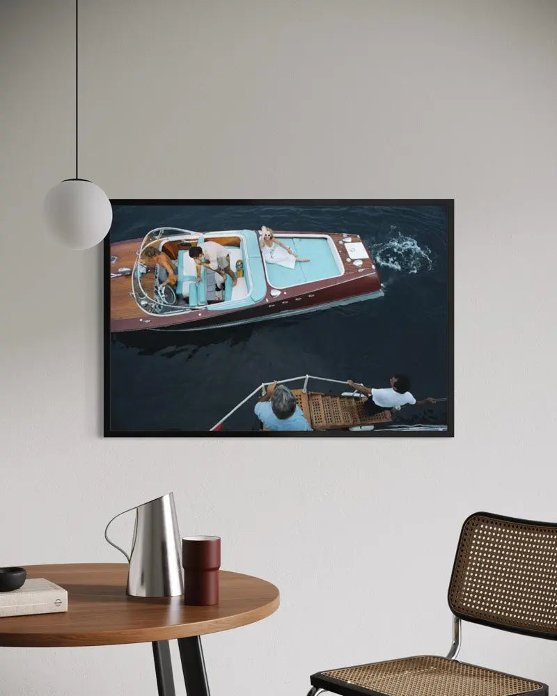 A framed art print depicting an aerial view of a wooden motorboat on dark water, displayed on a light gray wall in a modern room setting with a wooden table, chair, and pendant light. The Picturalist Wall Art and Photography for Designed Interiors. Slim Aarons Collection, Riva Boat. Getty Images Gallery.