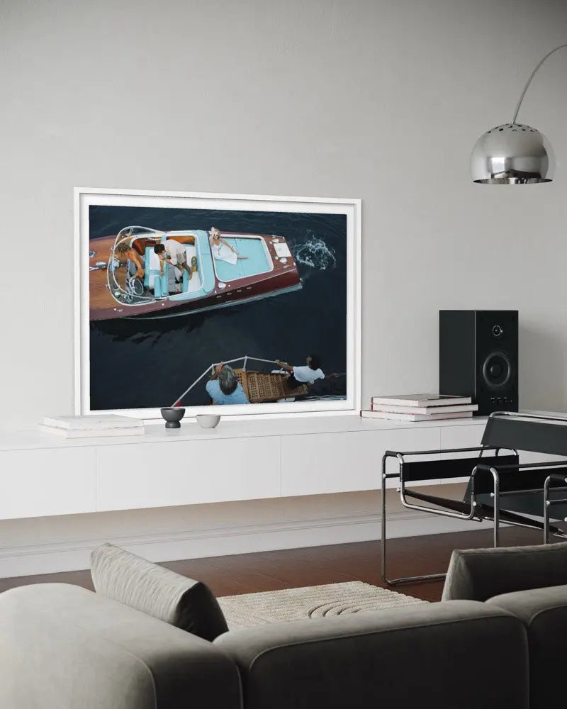 A framed art print depicting a classic motorboat on dark water, displayed on a white wall in a modern living room setting with a gray sofa, chair, and floor lamp. The Picturalist Wall Art and Photography for Designed Interiors. Slim Aarons Collection, Riva Boat. Getty Images Gallery.