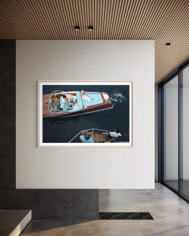 A framed art print depicting an aerial view of a red and blue motorboat on dark water, displayed on a white wall in a modern interior setting with a slatted wood ceiling. The Picturalist Wall Art and Photography for Designed Interiors. Slim Aarons Collection, Riva Boat. Getty Images Gallery.