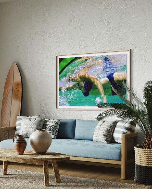 A framed square art print depicting a swimmer in turquoise water, displayed on a light gray wall above a blue sofa in a styled living room. The Picturalist Wall Art and Photography for Designed Interiors. Slim Aarons Collection, Poolside. Getty Images Gallery.