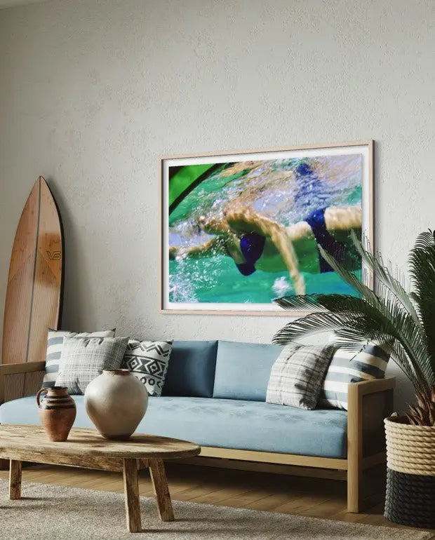 A framed square art print depicting a swimmer in turquoise water, displayed on a light gray wall above a blue sofa in a styled living room. The Picturalist Wall Art and Photography for Designed Interiors. Slim Aarons Collection, Poolside. Getty Images Gallery.