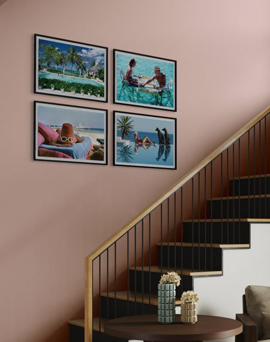 Four framed tropical beach and pool lifestyle photographs arranged in a 2x2 grid on a dusty pink wall above a staircase. The Picturalist Wall Art and Photography for Designed Interiors. Slim Aarons Collection, Poolside. Getty Images Gallery.