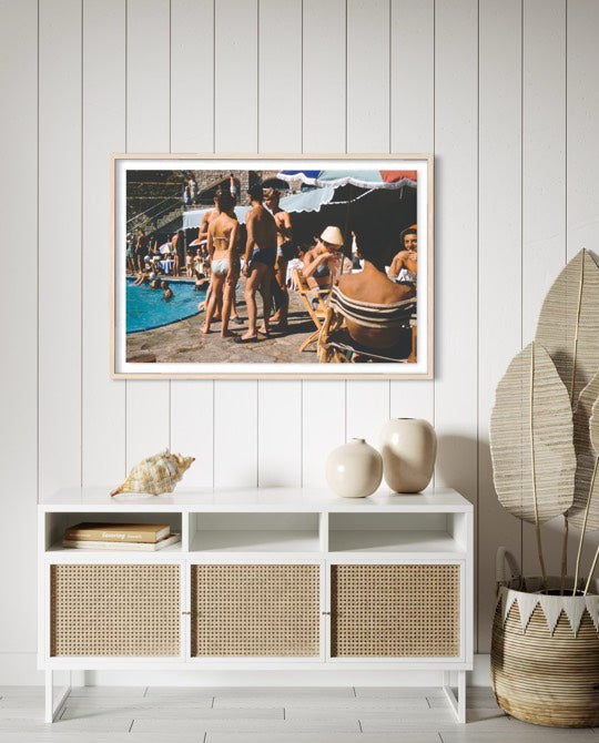 A framed art print depicting a vintage beach pool party scene, styled on a white paneled wall above a white sideboard cabinet with woven cane doors, flanked by decorative ceramic vases and a potted plant. The Picturalist Wall Art and Photography for Designed Interiors. Slim Aarons Collection, Poolside. Getty Images Gallery.