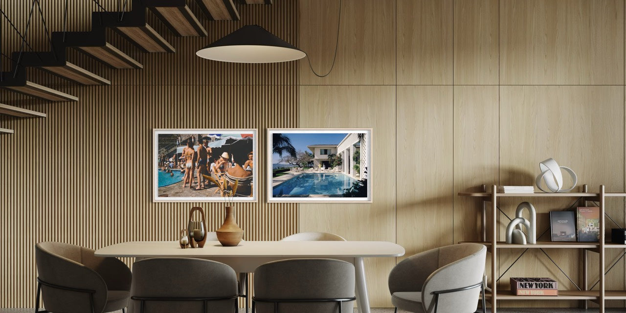 Two framed art prints depicting poolside scenes, displayed side-by-side on a light wood paneled wall in a modern dining room setting. The Picturalist Wall Art and Photography for Designed Interiors. Slim Aarons Collection, Poolside. Getty Images Gallery.