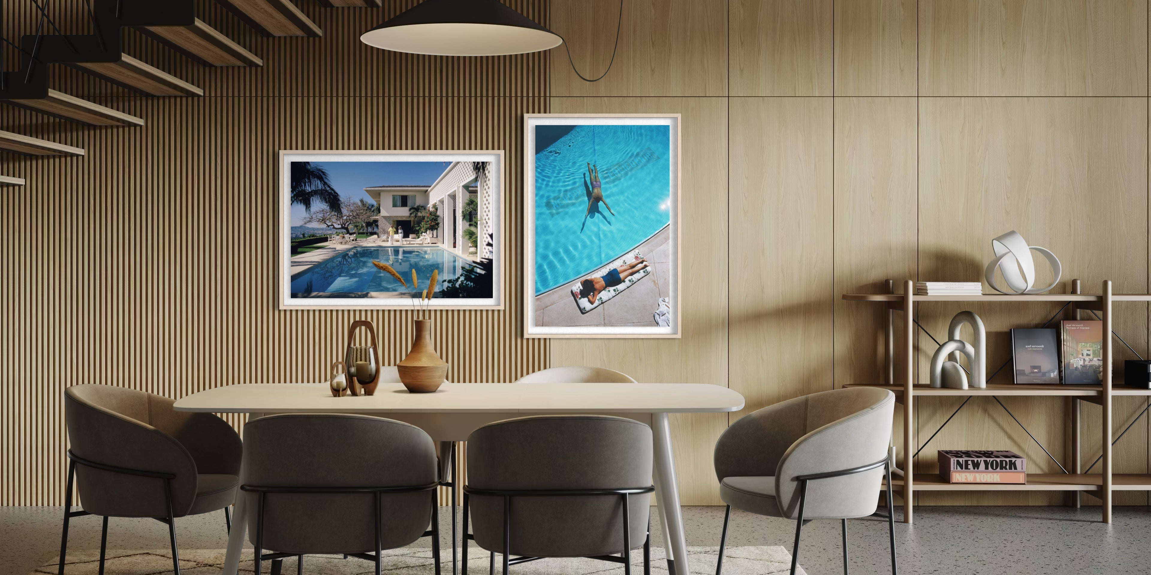 Two framed art prints hanging on a light wood slatted wall above a dining table. The left print shows a poolside villa scene, and the right print shows an aerial view of a swimmer in a bright blue pool. The Picturalist Wall Art and Photography for Designed Interiors. Slim Aarons Collection, Poolside. Getty Images Gallery.