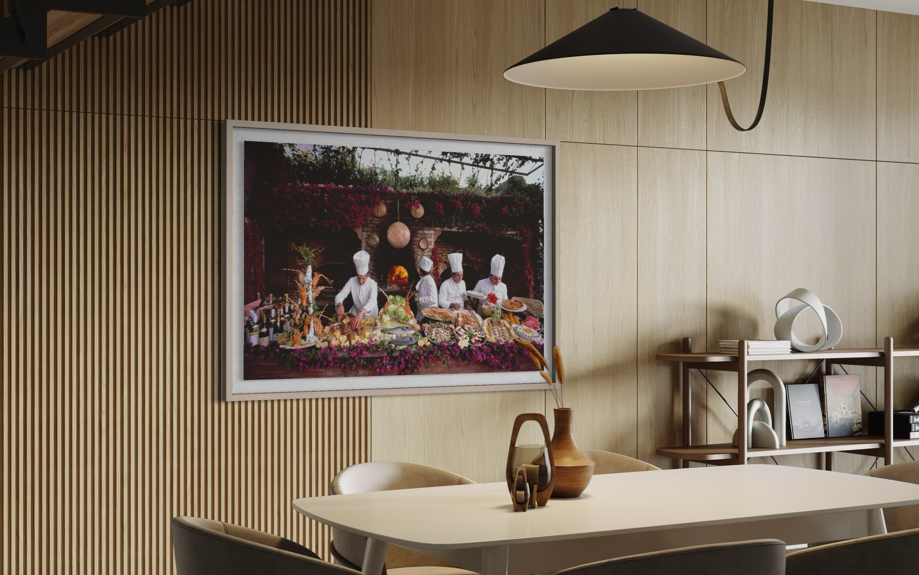A framed art print depicting a group of chefs preparing a lavish meal, displayed on a light wood slatted wall in a modern dining room setting. The Picturalist Wall Art and Photography for Designed Interiors. Slim Aarons Collection, Poolside. Getty Images Gallery.