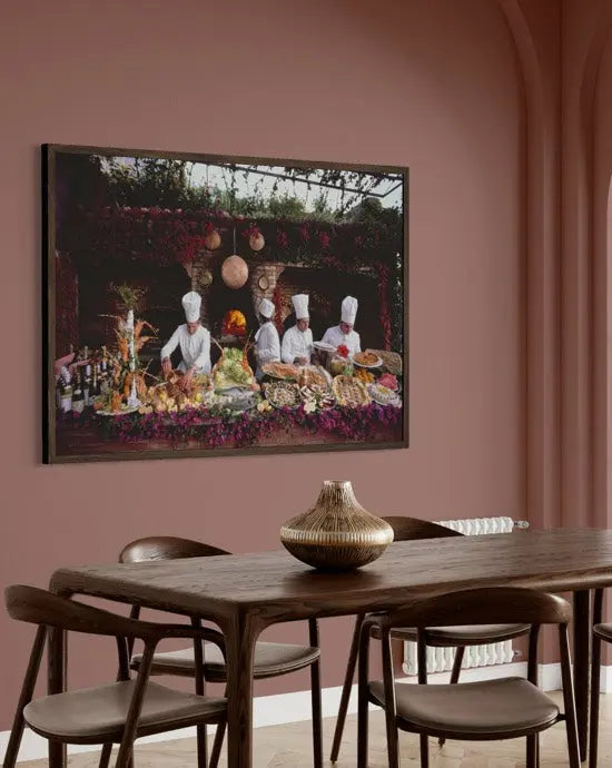 A framed art print depicting chefs serving food hangs on a dusty rose wall above a wooden dining table with chairs. The Picturalist Wall Art and Photography for Designed Interiors. Slim Aarons Collection, Poolside. Getty Images Gallery.
