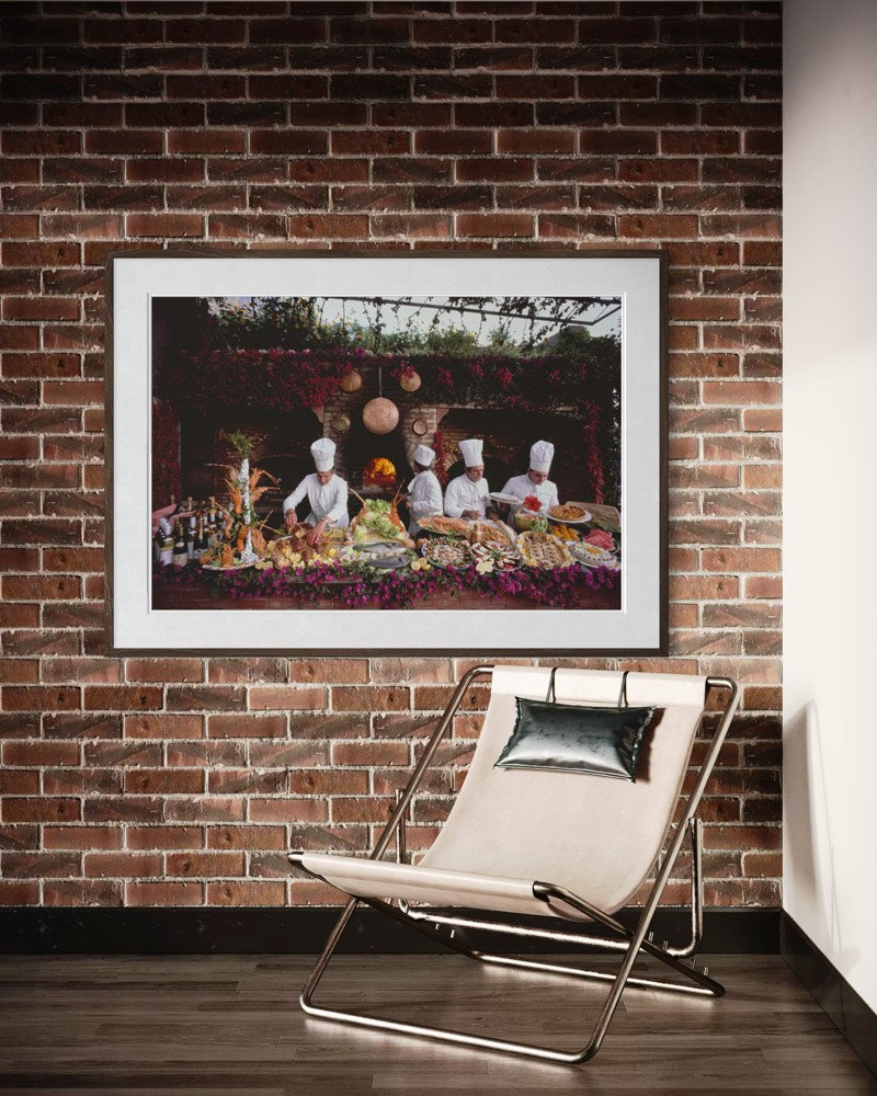 A framed art print depicting a restaurant scene with chefs and food, displayed on a brick wall above a modern beige lounge chair. The Picturalist Wall Art and Photography for Designed Interiors. Slim Aarons Collection, Poolside. Getty Images Gallery.