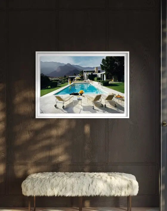 A framed art print depicting a luxurious outdoor poolside scene with mountains in the background, displayed on a dark wood-paneled wall above a white faux-fur bench. The Picturalist Wall Art and Photography for Designed Interiors. Slim Aarons Collection, Poolside. Getty Images Gallery.