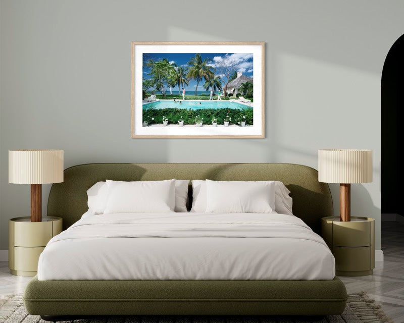 A framed tropical beach print with palm trees, a swimming pool, and blue sky, displayed on a light gray wall above a modern bed in a styled bedroom. The Picturalist Wall Art and Photography for Designed Interiors. Slim Aarons Collection, Poolside. Getty Images Gallery.