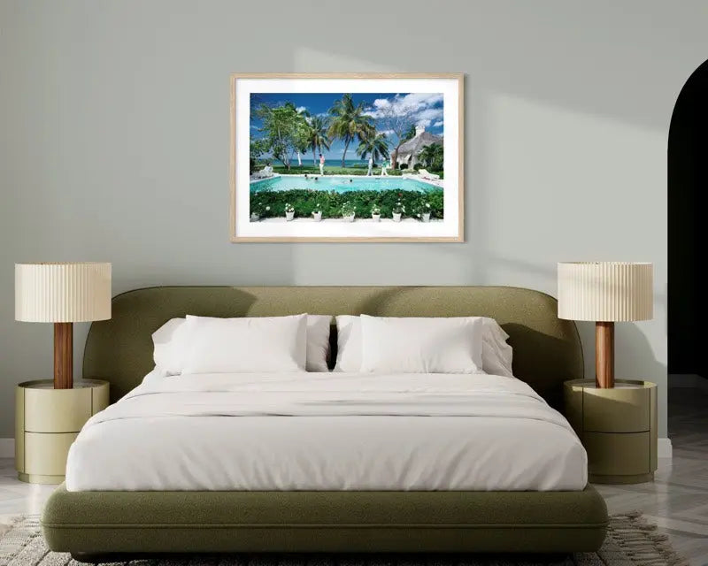 A framed tropical beach print with palm trees, a swimming pool, and blue sky, displayed on a light gray wall above a modern bed in a styled bedroom. The Picturalist Wall Art and Photography for Designed Interiors. Slim Aarons Collection, Poolside. Getty Images Gallery.