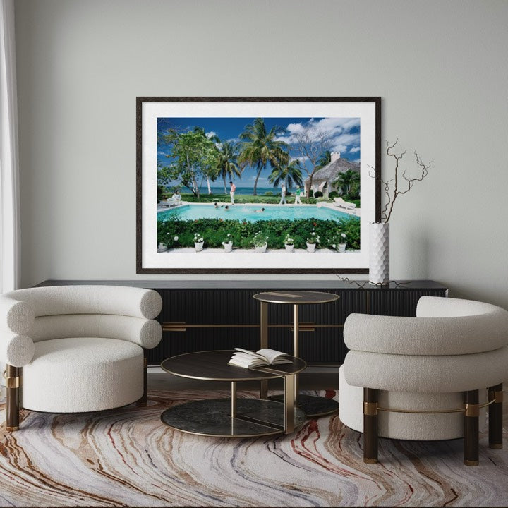 A framed art print depicting a tropical beach resort with palm trees, a swimming pool, and a thatched-roof structure, displayed on a light gray wall above a black console table in a modern living room setting. The Picturalist Wall Art and Photography for Designed Interiors. Slim Aarons Collection, Poolside. Getty Images Gallery.