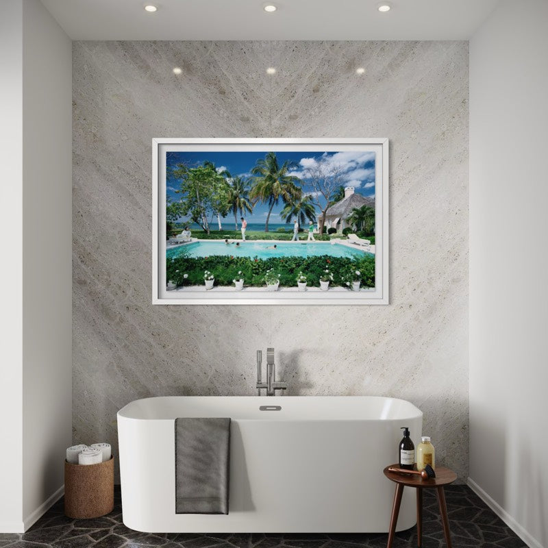 A framed wall art print depicting a tropical beach resort pool scene with palm trees, blue sky, and white umbrellas, displayed above a freestanding white bathtub in a modern bathroom. The Picturalist Wall Art and Photography for Designed Interiors. Slim Aarons Collection, Poolside. Getty Images Gallery.