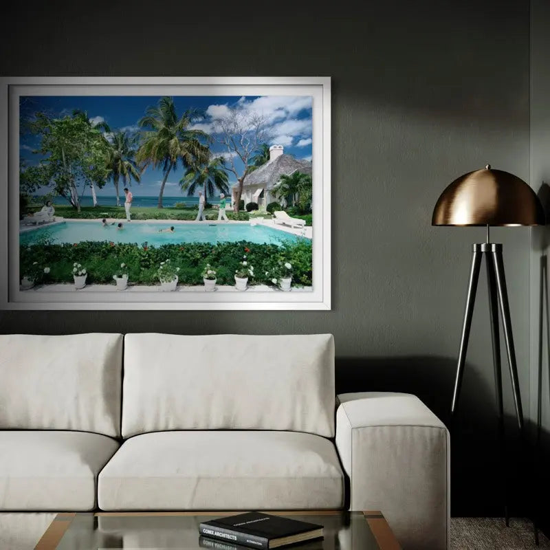 A large framed tropical resort landscape print hangs on a dark gray wall above a cream-colored sofa. The artwork depicts palm trees, a swimming pool, beach, and a thatched-roof building. The Picturalist Wall Art and Photography for Designed Interiors. Slim Aarons Collection, Poolside. Getty Images Gallery.