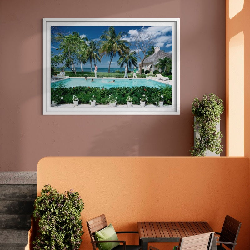 A framed landscape art print depicting a tropical resort pool scene with palm trees, lounge chairs, and a thatched-roof building, displayed on a terracotta wall above an outdoor patio with a table and chairs. The Picturalist Wall Art and Photography for Designed Interiors. Slim Aarons Collection, Poolside. Getty Images Gallery.