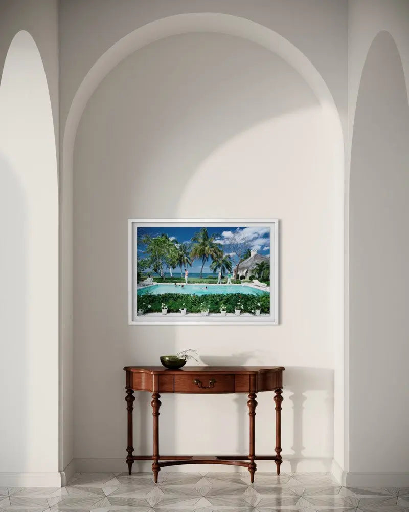 A framed tropical beach and poolside landscape print hangs on a white wall in an arched interior setting above a wooden console table. The Picturalist Wall Art and Photography for Designed Interiors. Slim Aarons Collection, Poolside. Getty Images Gallery.