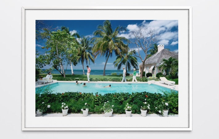 A framed art print depicting a tropical resort scene with a swimming pool, palm trees, ocean, and a thatched-roof building, shown against a light gray background. The Picturalist Wall Art and Photography for Designed Interiors. Slim Aarons Collection, Poolside. Getty Images Gallery.