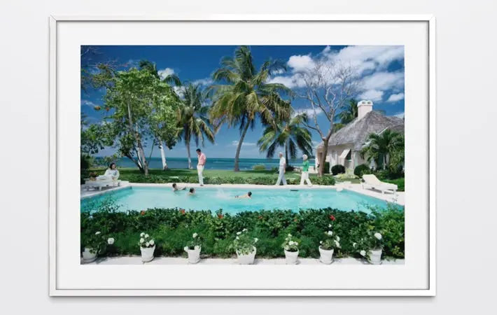 A framed art print depicting a tropical resort scene with a swimming pool, palm trees, ocean, and a thatched-roof building, shown against a light gray background. The Picturalist Wall Art and Photography for Designed Interiors. Slim Aarons Collection, Poolside. Getty Images Gallery.