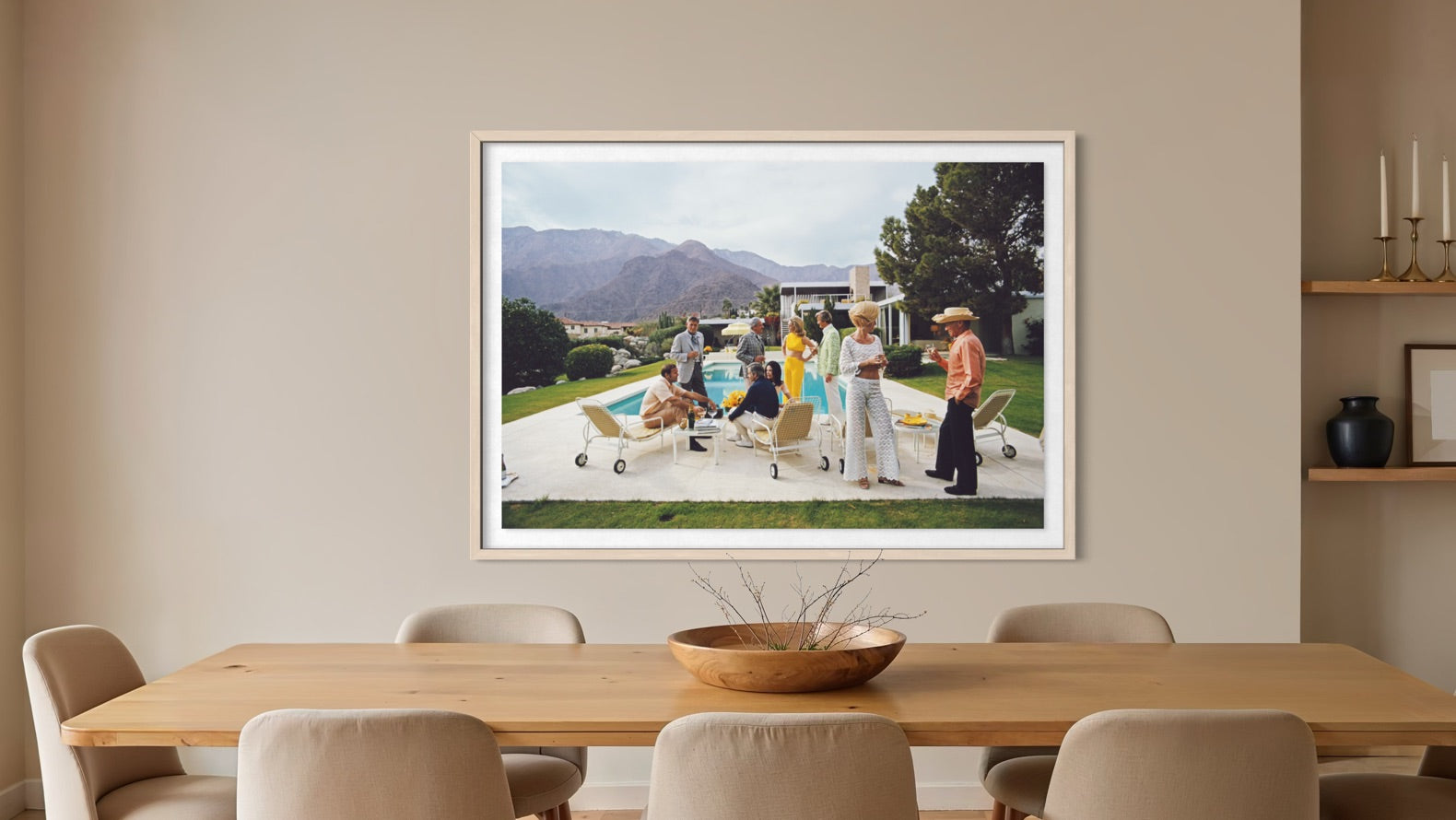 A framed art print depicting a group of people relaxing by a pool in a sunny outdoor setting, displayed on a beige wall above a wooden dining table in a modern dining room. The Picturalist Wall Art and Photography for Designed Interiors. Slim Aarons Collection, Poolside. Getty Images Gallery.
