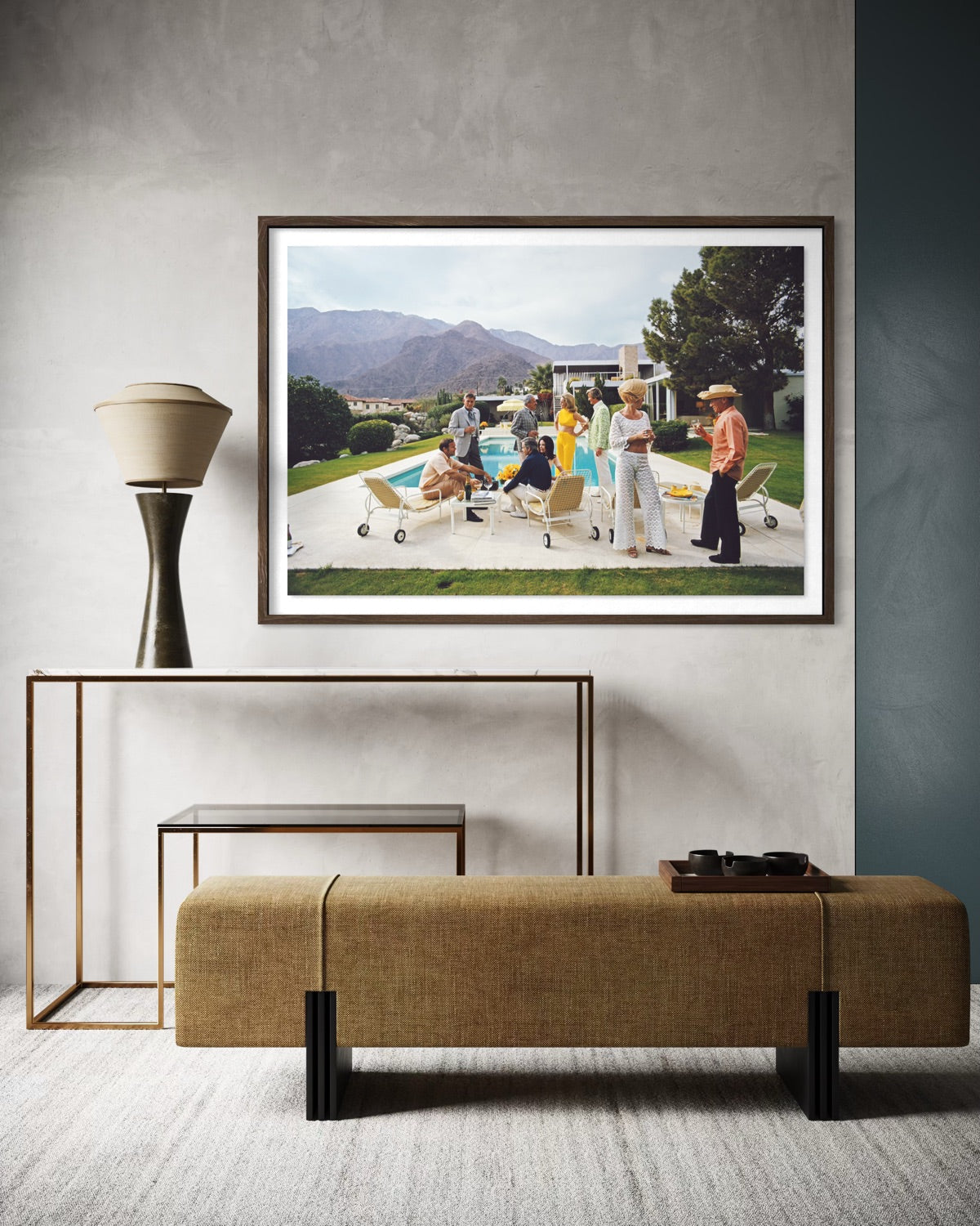 A framed art print depicting a vintage outdoor scene with people and a pool, styled in a modern interior setting above a textured bench and console table. The Picturalist Wall Art and Photography for Designed Interiors. Slim Aarons Collection, Poolside. Getty Images Gallery.