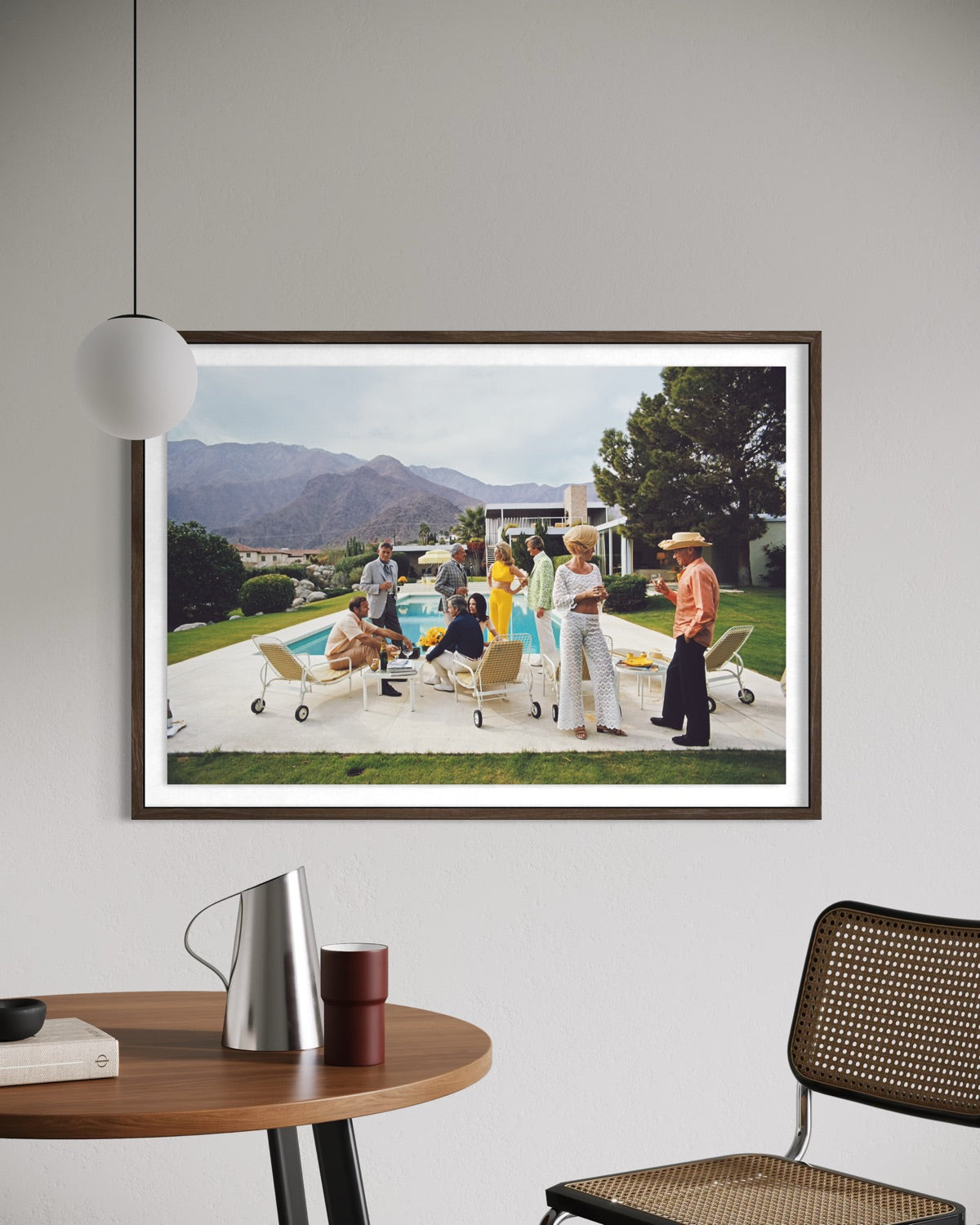 A framed art print depicting a 1960s-style pool party scene with people, mountains, and palm trees, displayed on a light gray wall above a wooden table with a silver pitcher and a woven chair. The Picturalist Wall Art and Photography for Designed Interiors. Slim Aarons Collection, Poolside. Getty Images Gallery.